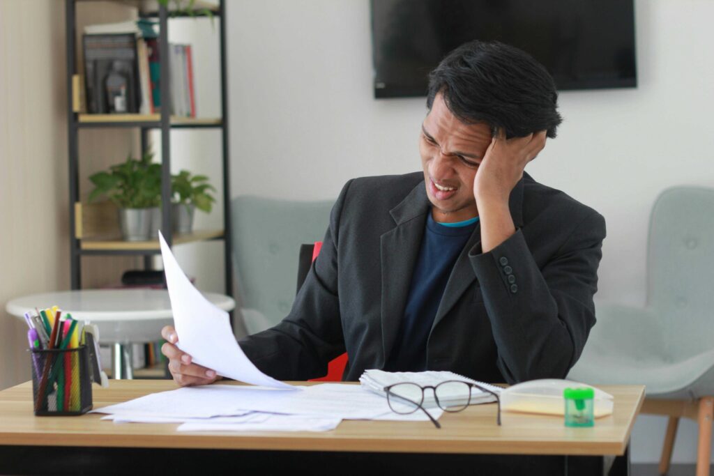 Professional man in a dark blazer sits at a desk, gripping papers with one hand while resting his head in the other, looking stressed in a bright office.