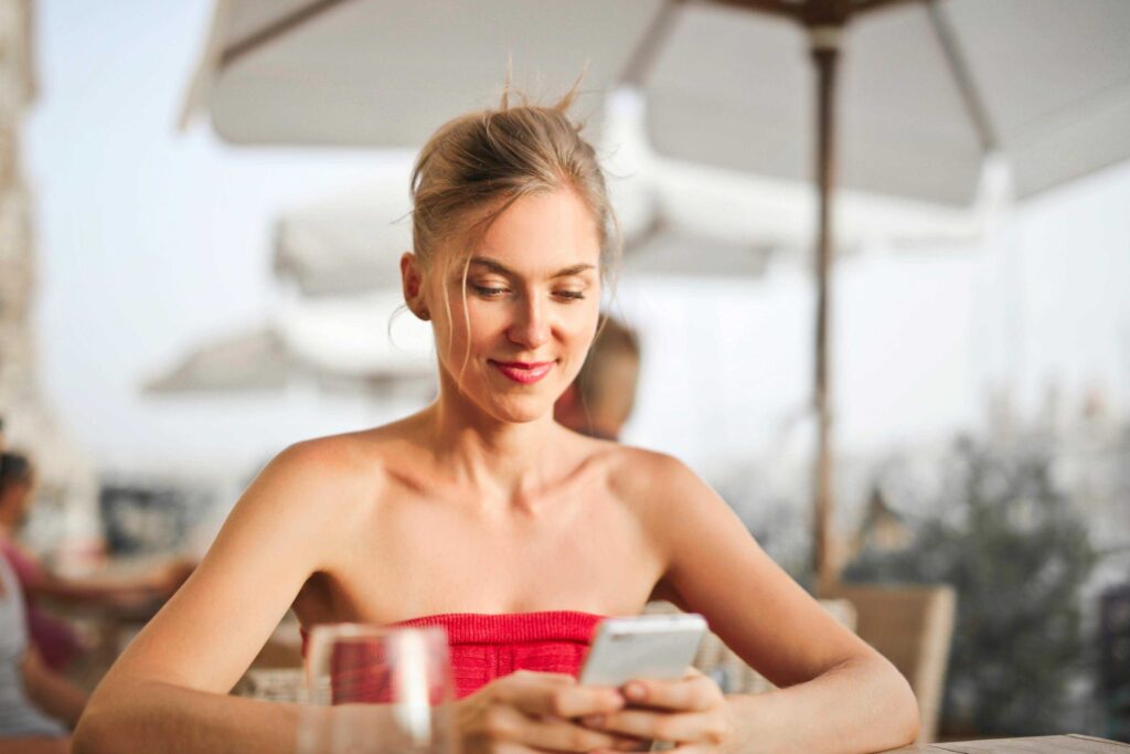 Woman in a red strapless top sits at an outdoor cafe, looking at her smartphone with a drink on the table in front of her.