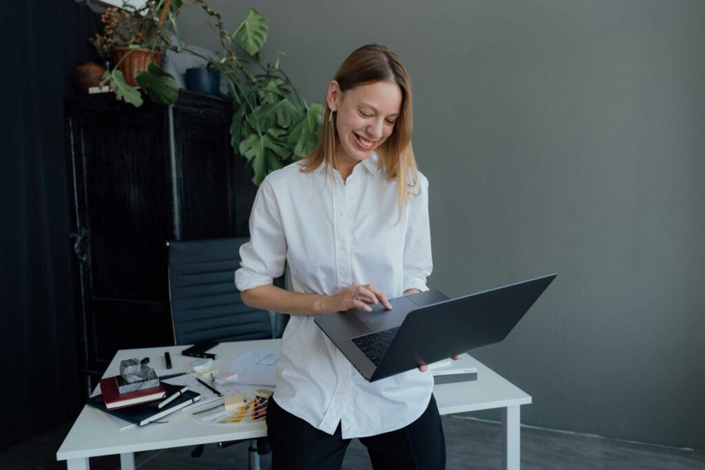 Smiling woman in a white shirt holds an open laptop at a desk with color swatches and papers nearby.