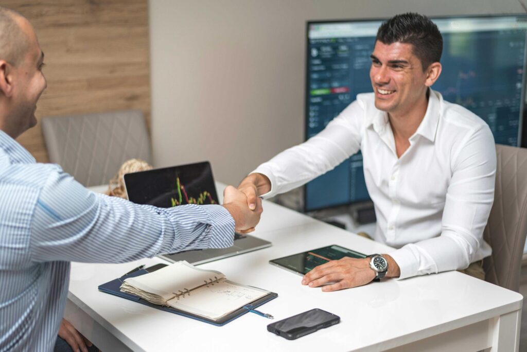 Two businessmen shake hands across a white desk in an office, with a laptop, notebook, and phone on the table and charts on a monitor in the background.