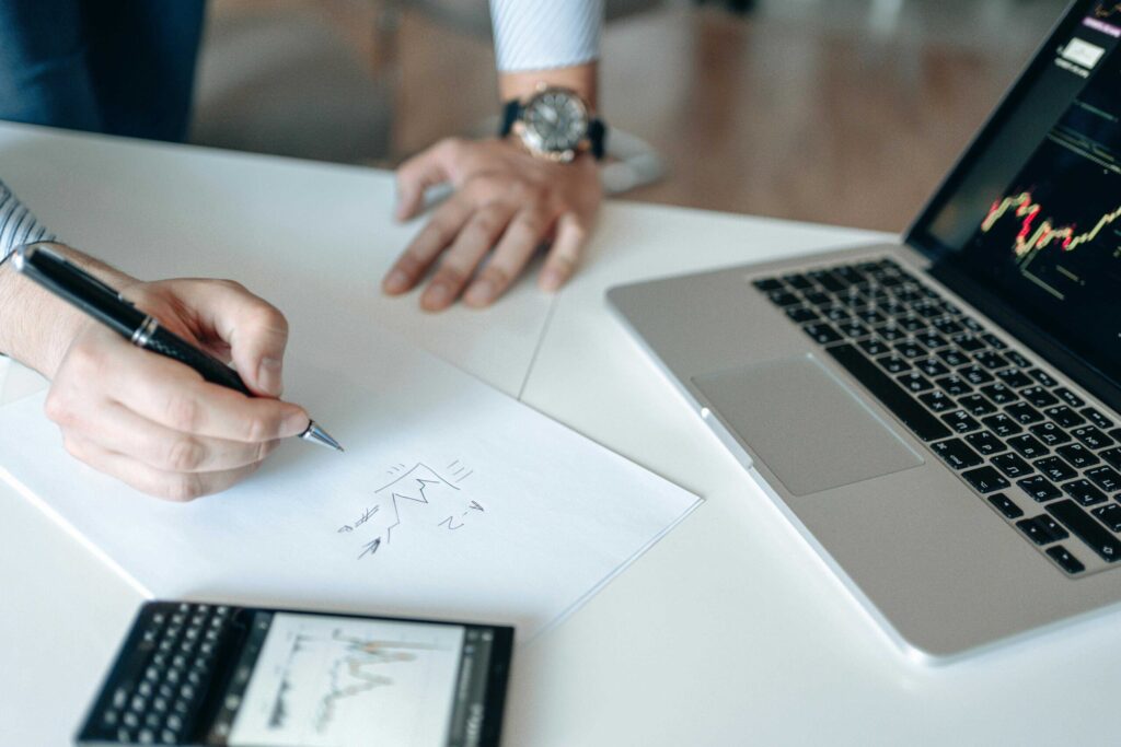 Two hands sketching on a sheet of paper while a person with a wristwatch rests a hand nearby, with a laptop showing a graph on the screen nearby.