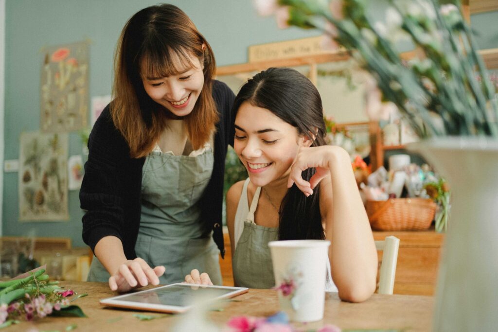 Two women in aprons collaborate on a crafting project at a wooden table with flowers and a tablet nearby, in a bright workshop of greenery and supplies.