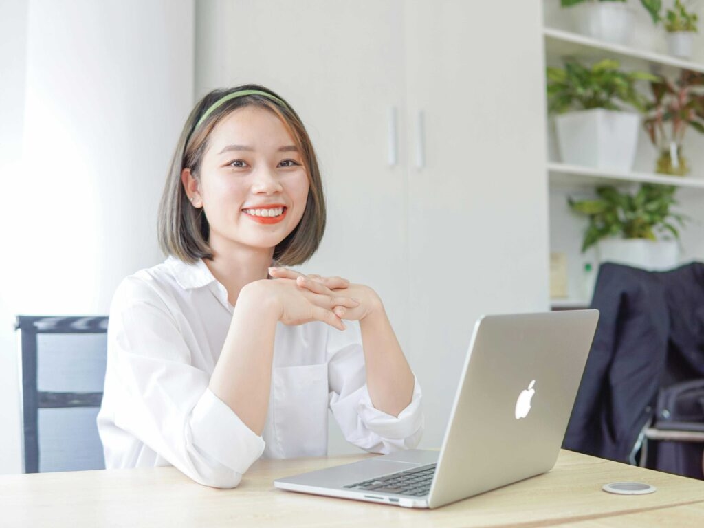 Smiling woman in a white shirt sits at a desk with an open laptop in a bright office setting with plants in the background.