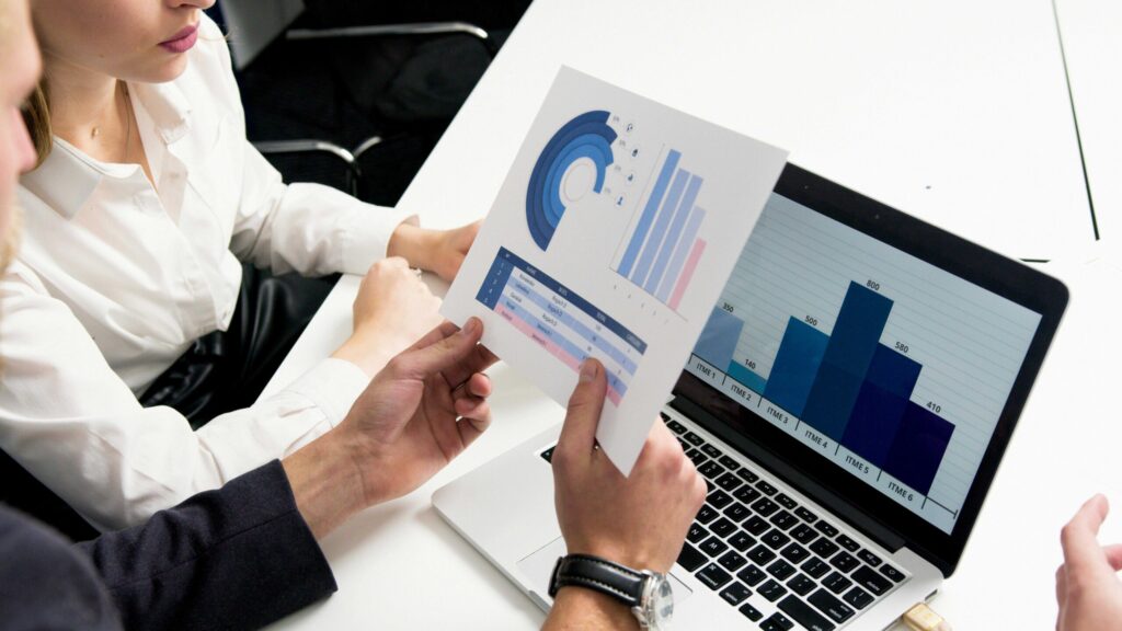 Team members review printed charts beside a laptop displaying bar graphs and a pie chart.