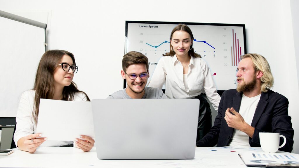 Four colleagues gathered around a laptop in a bright office, discussing charts displayed on a screen behind them.
