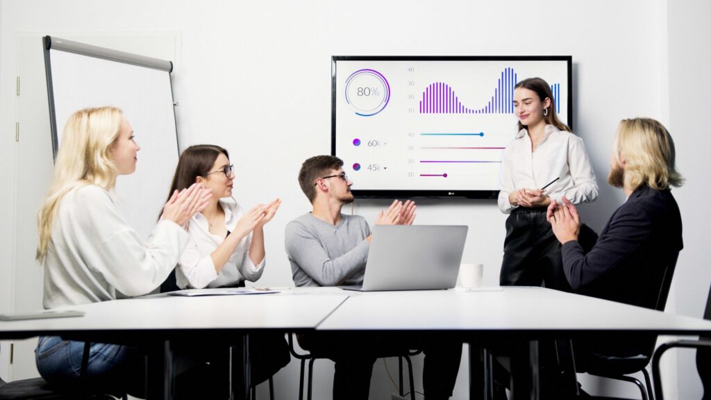 Team of five applauding a presenter beside a large chart-filled screen in a conference room.