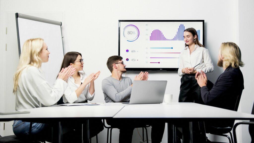 Five colleagues in a conference room applaud as a presenter explains graphs on a large screen.