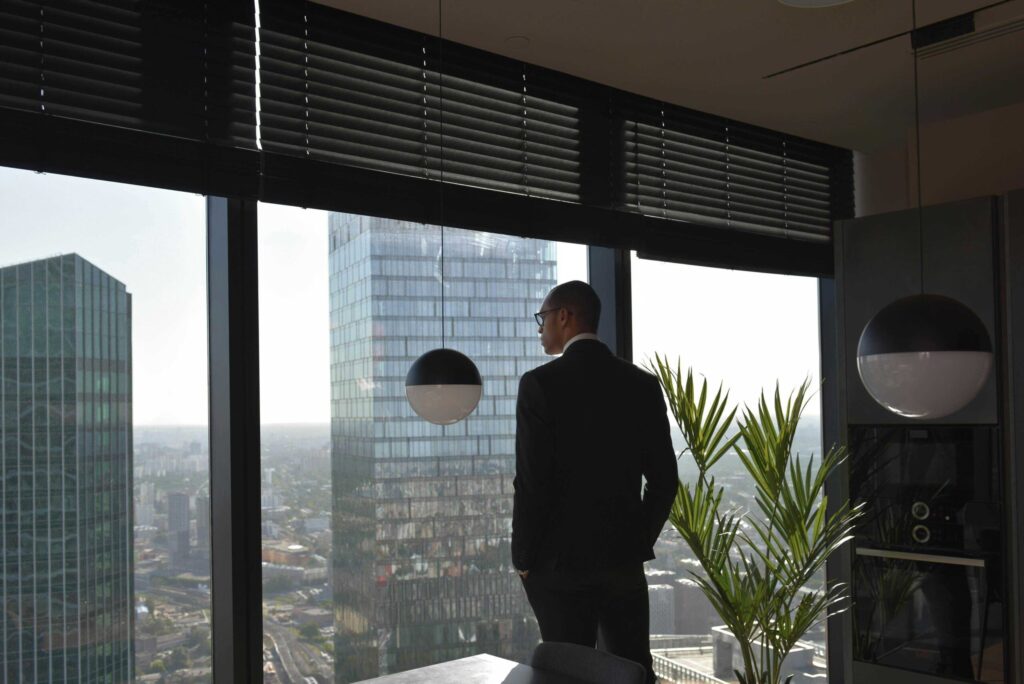 Businessman in a suit stands by a window with blinds, overlooking a city skyline.