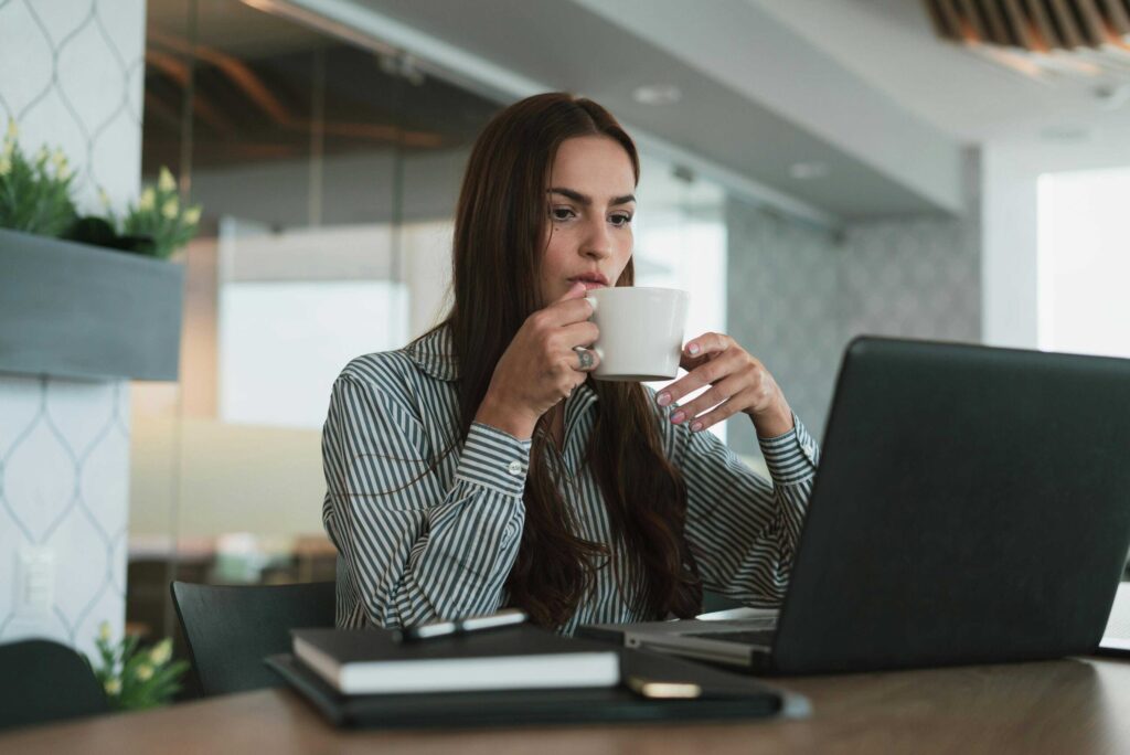 Woman in a striped shirt sipping from a white mug at a desk with a laptop in a modern office setting