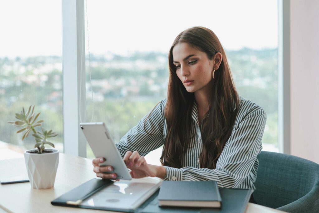 Woman in a striped shirt sits at a bright desk by a window, looking at her smartphone with a plant and notebooks nearby.