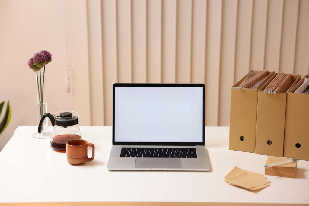 Laptop centered on a white desk with a blank screen, flanked by a glass coffee pot and an orange mug; purple bloom vase and brown file boxes in background.