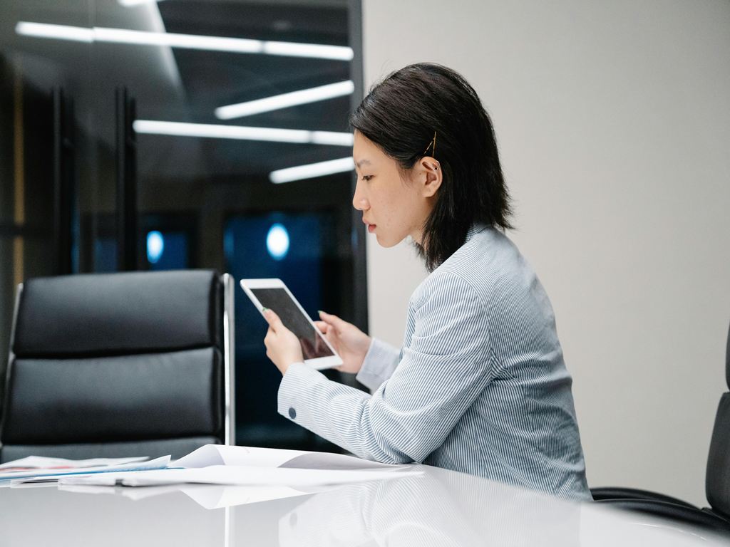 Businesswoman in a striped blazer sits at a conference table, looking at a tablet while reviewing documents