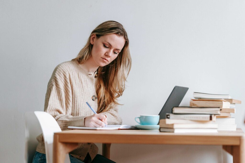 Woman writing at a wooden desk with a notebook, laptop, and a stack of books nearby in a bright room.