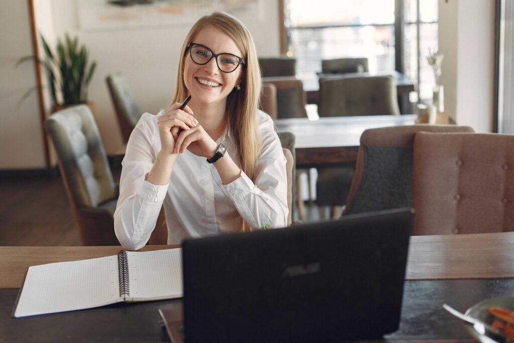 Smiling woman with glasses seated at a desk in a bright office, holding a pen beside an open notebook and laptop
