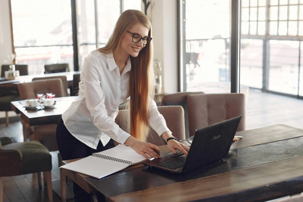 Woman with long hair and glasses working at a cafe, writing in a notebook while using a laptop.