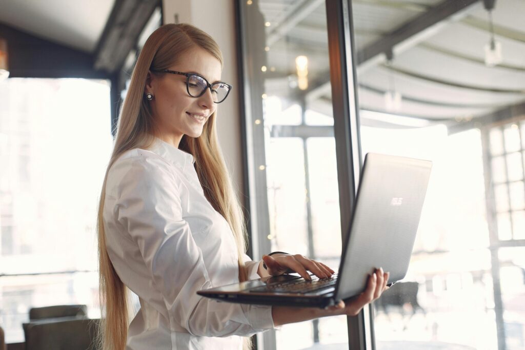 Woman with glasses wearing a white shirt, typing on a laptop by a large window in a bright office space