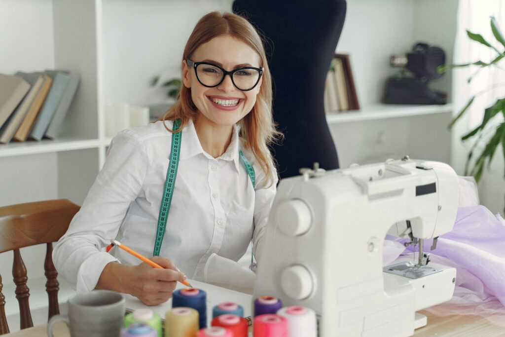 Smiling woman sewing at a machine with a turquoise measuring tape around her neck in a bright sewing room.