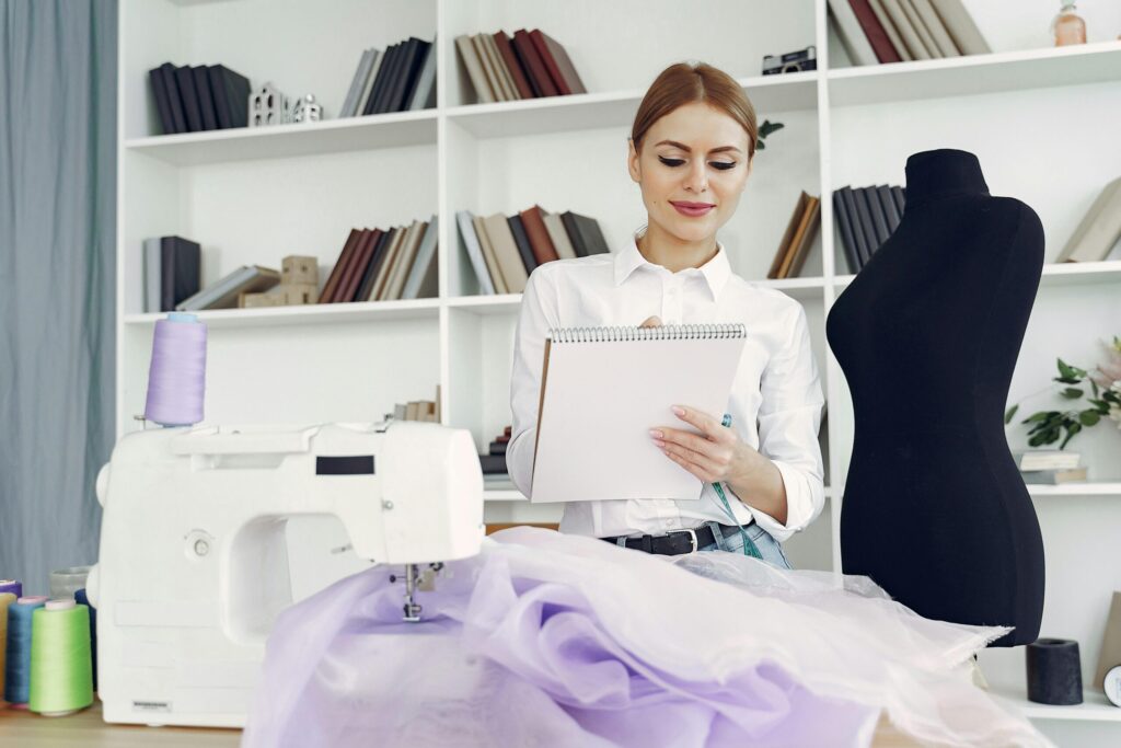 A woman in a white shirt holding a spiral notebook standing beside a sewing machine, examining lavender fabric on a worktable in a sewing studio with shelves and rolls of thread nearby.
