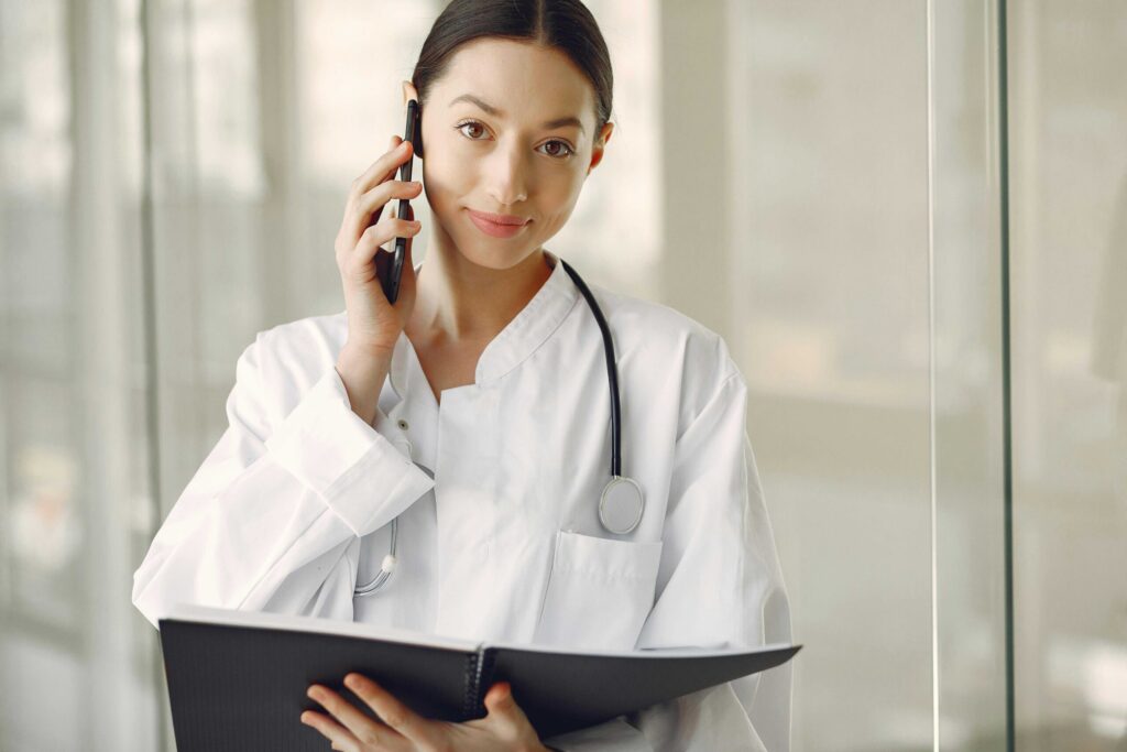 Female doctor in a white coat talks on a cellphone while holding a binder and wearing a stethoscope around her neck in a medical setting.