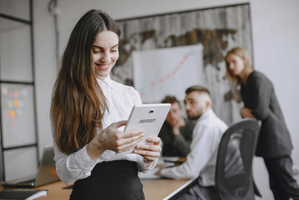 Smiling woman in a white blouse holds a tablet in a modern office with colleagues in the background.