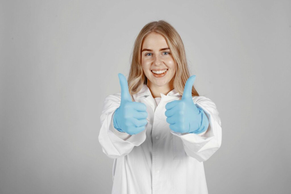 Smiling person in a white lab coat and blue gloves giving a thumbs-up toward the camera.