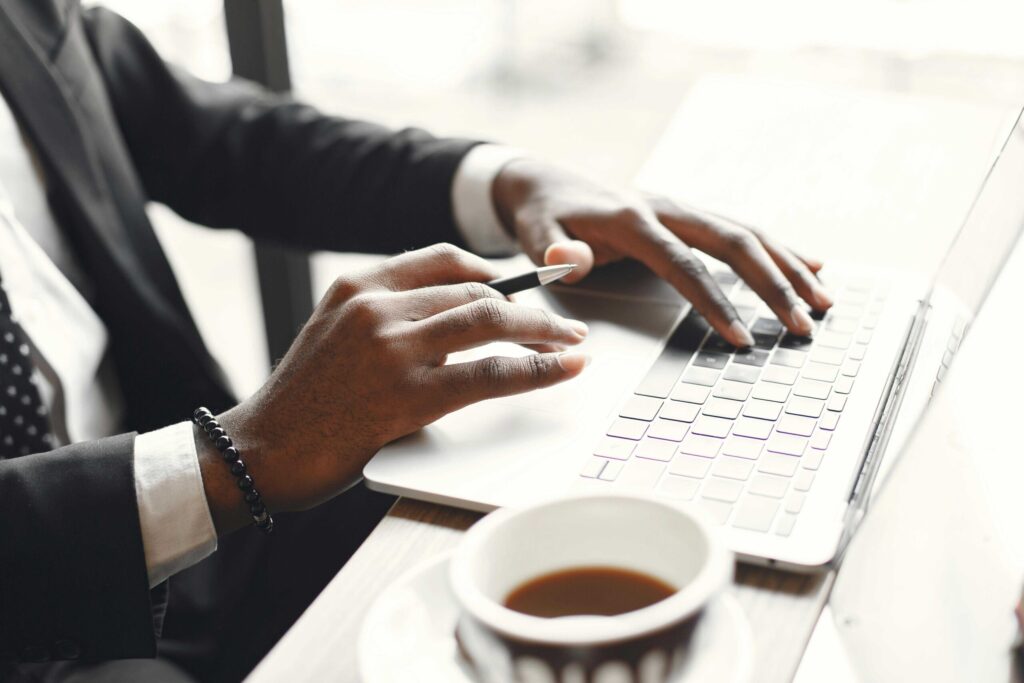 Person in a suit typing on a laptop with a pen in hand, coffee cup on a desk nearby.