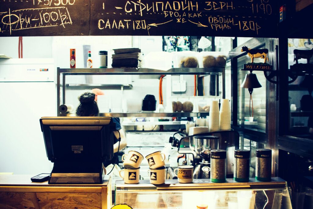 Cafe counter interior with a barista at the register, mugs stacked on the counter and a chalkboard menu overhead.