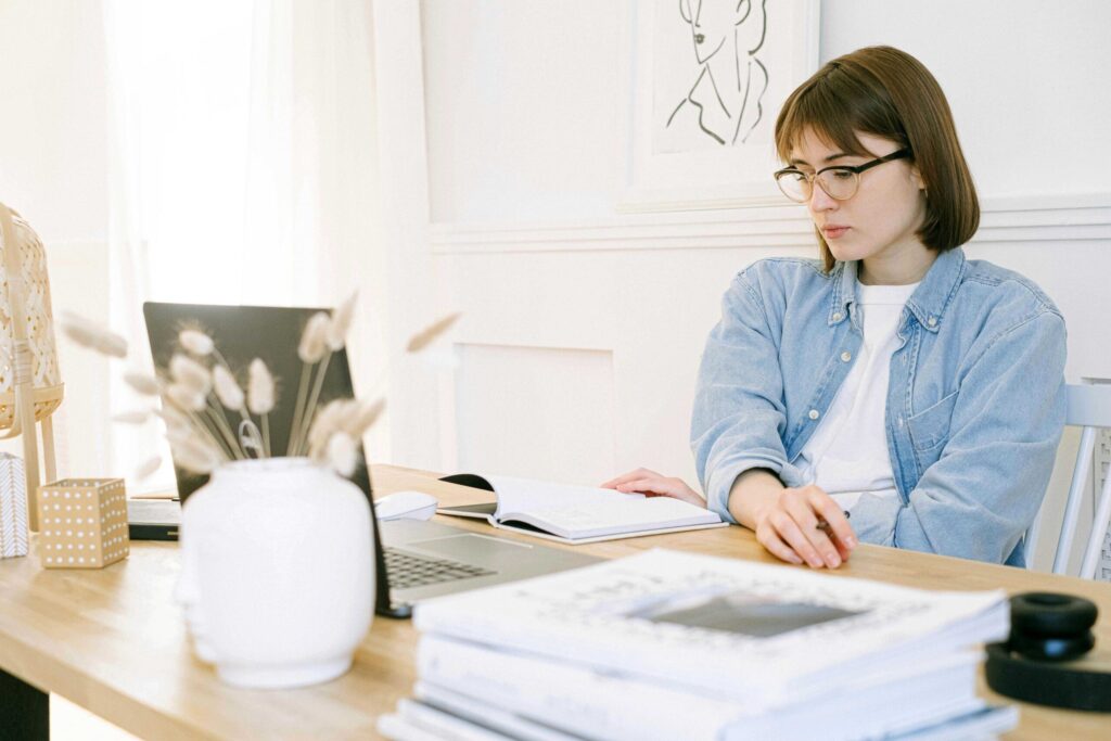 Woman with glasses working at a desk, studying with an open notebook and laptop in a bright, tidy room