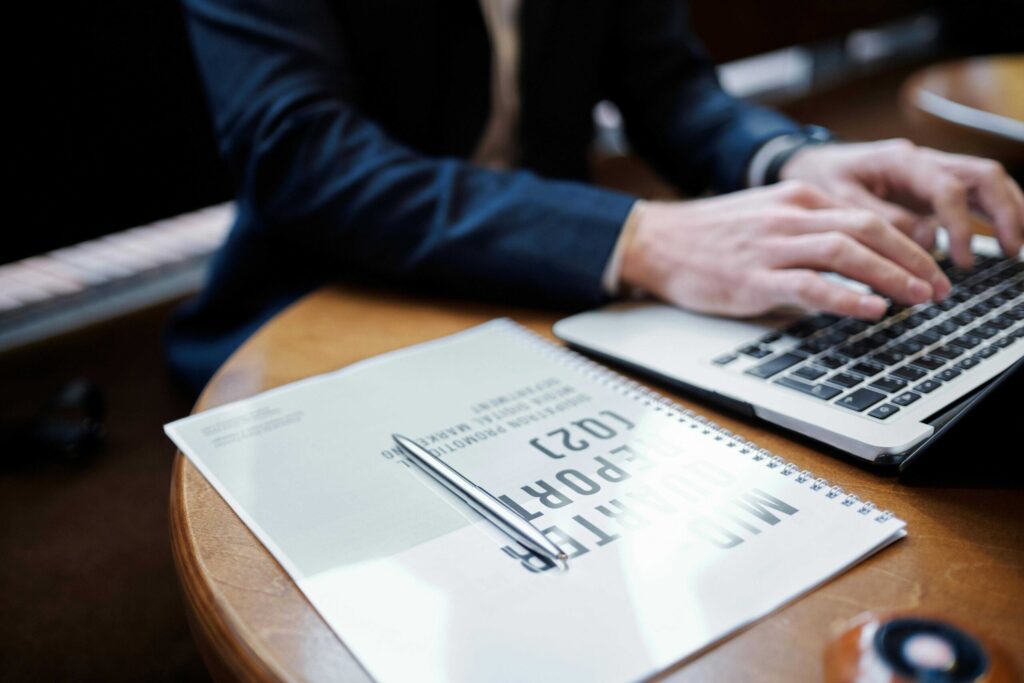 Hands typing on a laptop keyboard at a wooden desk with a spiral notebook nearby.