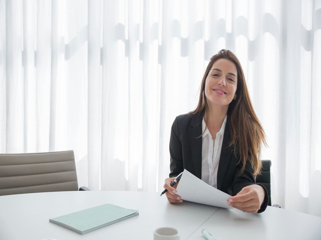Smiling professional woman in a black blazer sits at a white conference table, reviewing documents.