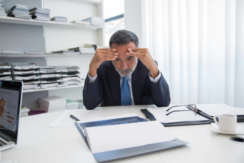 A stressed-looking man in a dark suit and blue tie sits at a desk with papers scattered around, hands pressed to his forehead.