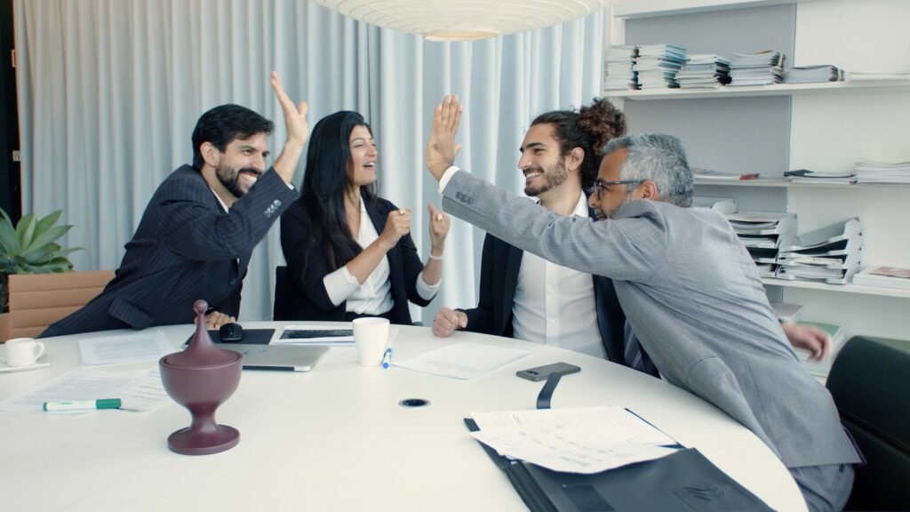 Four colleagues in a meeting room celebrate with high fives around a conference table.