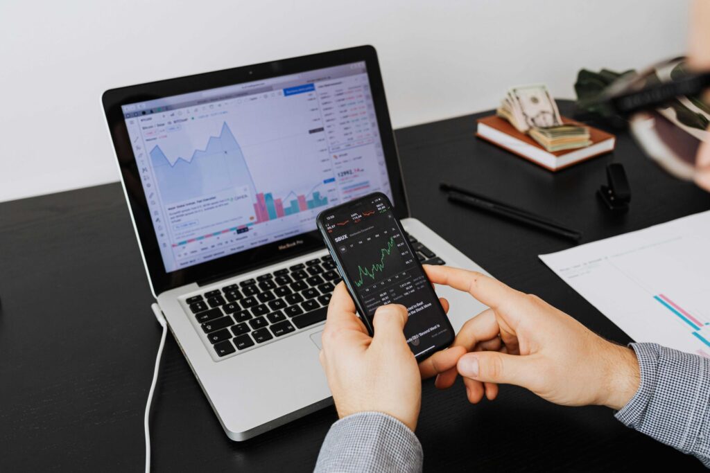 Person holding a smartphone displaying stock charts while a laptop shows financial graphs on a desk.