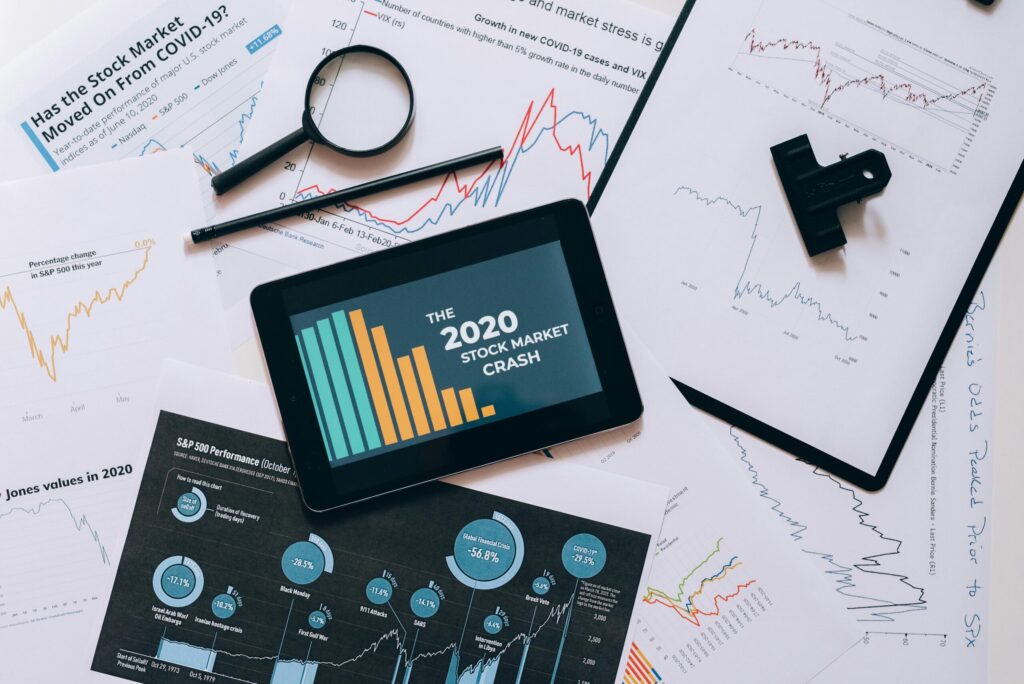 Tablet showing a report titled 'The 2020 Stock Market Crash' overlaid with bar charts and graphs amid scattered stock sheets and a magnifying glass