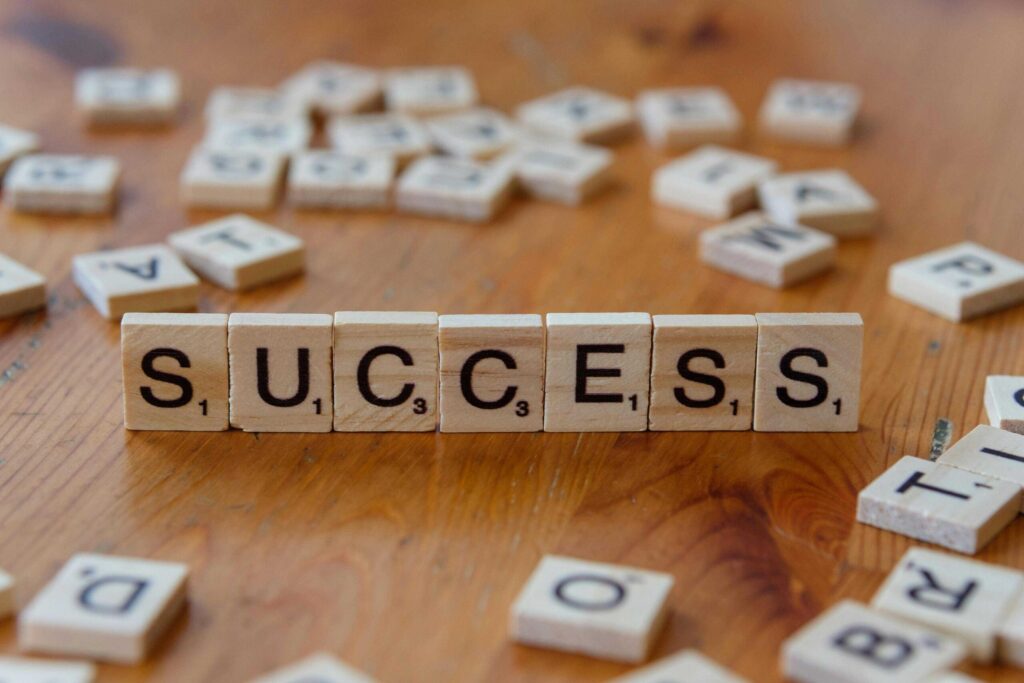 Scrabble tiles spelling the word SUCCESS on a wooden table, with letters and tile edges visible and scattered tiles in the background.