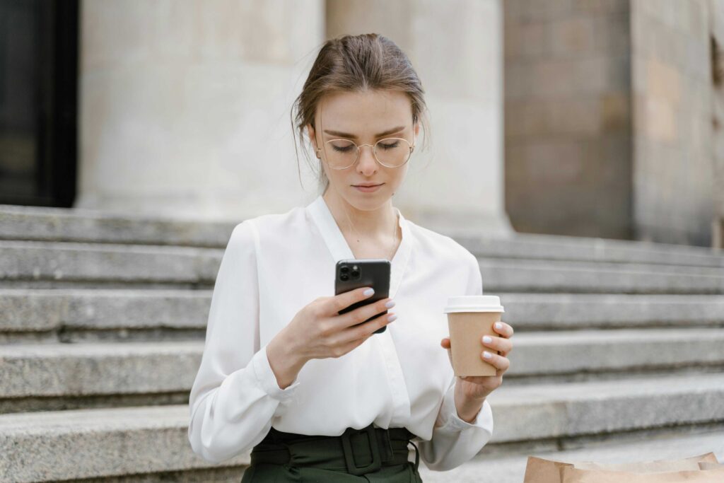 Woman seated on outdoor steps, looking at her smartphone while holding a paper coffee cup; wearing a white blouse and round glasses.