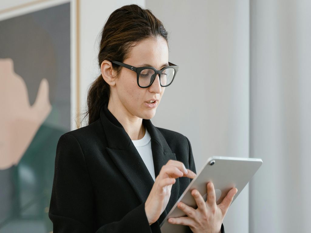 Professional woman in a blazer wearing glasses, using a tablet in an office setting by a window.