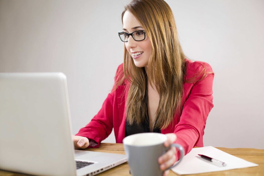 Woman wearing glasses works on a laptop at a desk, holding a coffee mug and smiling
