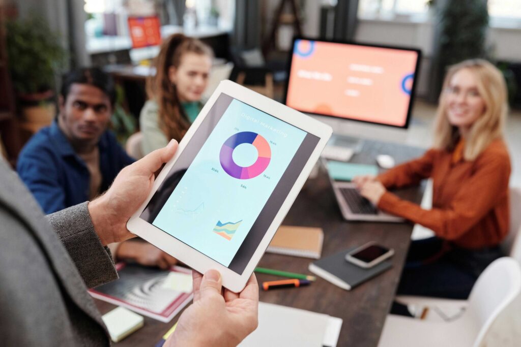 Person holding a tablet displaying a colorful pie chart and graphs to a group in a meeting room
