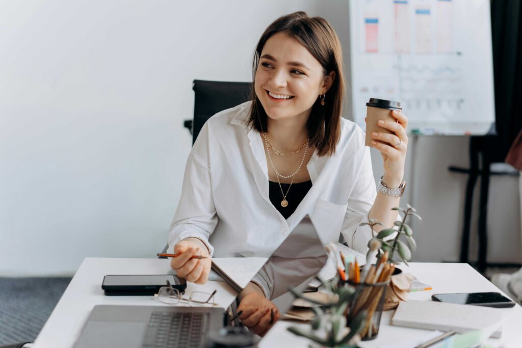 Smiling woman in a white blouse sits at a desk, holding a takeaway coffee cup with office supplies and a laptop nearby.