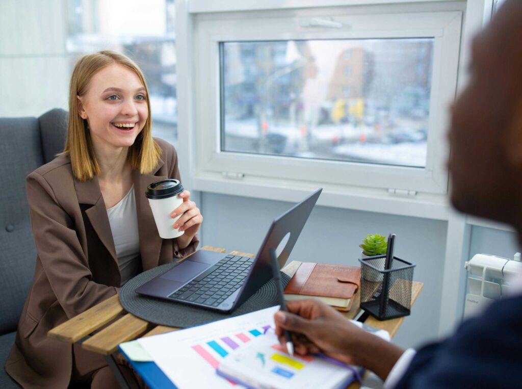 Smiling woman in a brown blazer holds a coffee cup during a meeting at a laptop-filled desk by a window.