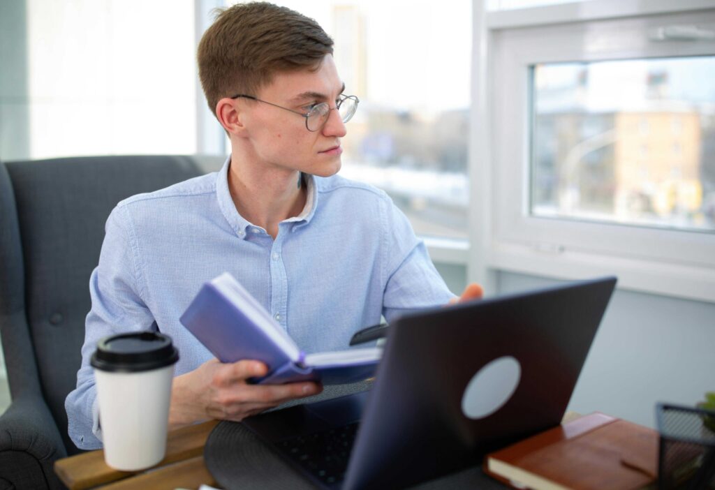 Man wearing glasses in a light blue shirt sits at a desk with a laptop and a purple notebook in hand inside a bright office.