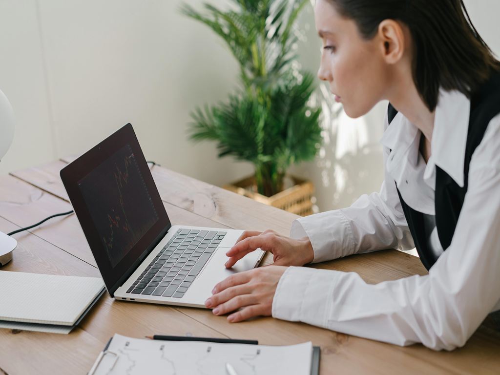 Businesswoman in a white shirt and black vest types on a laptop at a wooden desk; screen shows financial charts.