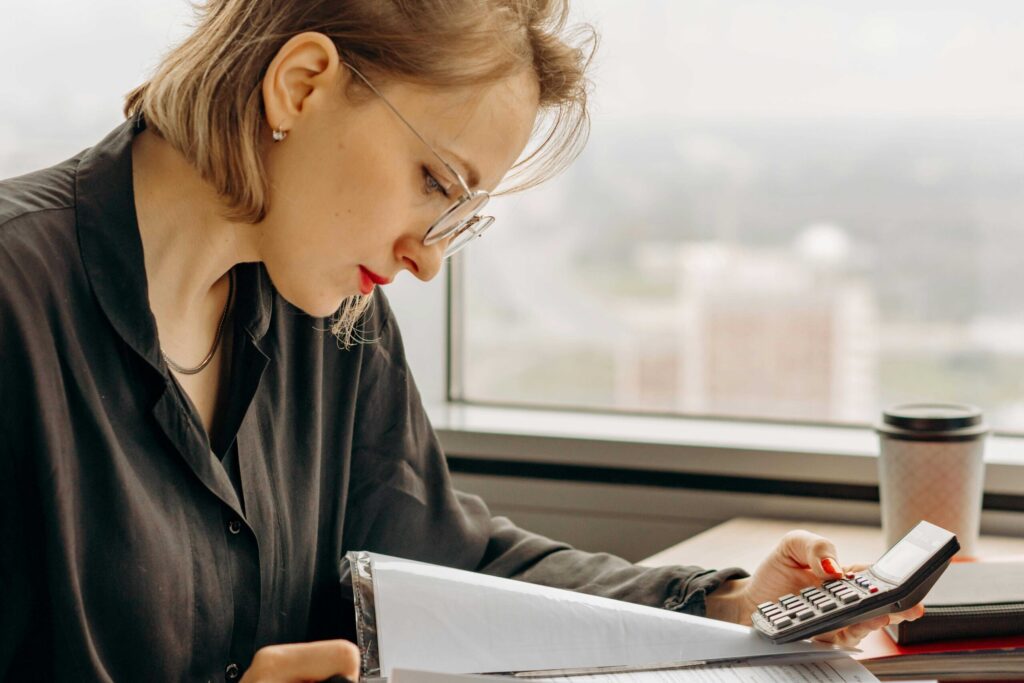 Woman in a dark shirt uses a calculator and reviews documents at a desk by a window, with a coffee cup nearby.
