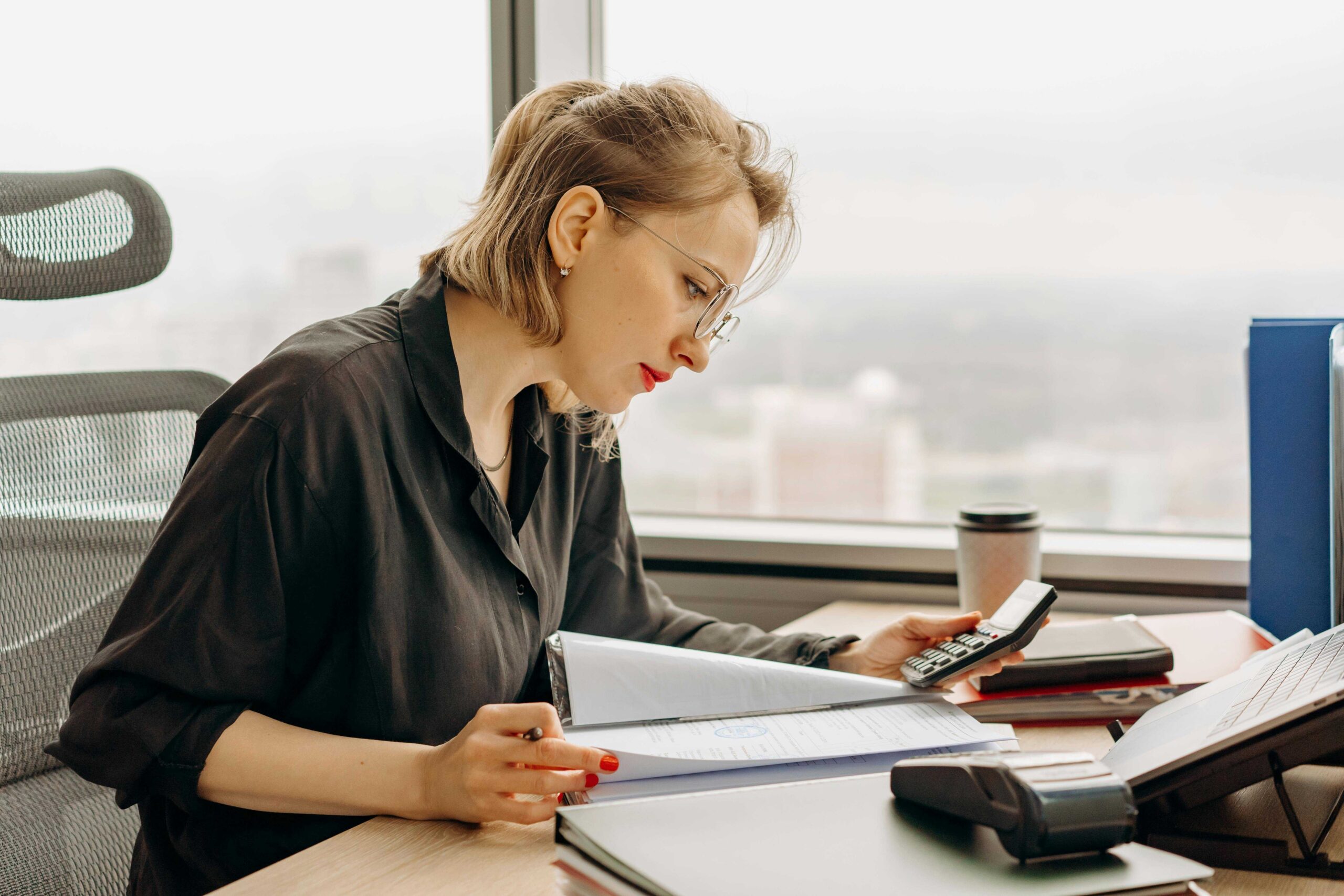 Professional woman in a black shirt reviewing documents at a desk, with a calculator and coffee cup nearby in a bright office.