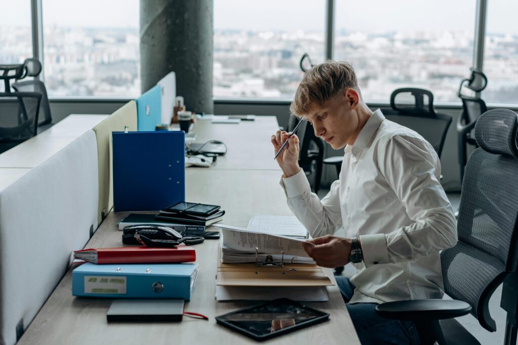 Man in a white shirt sits at a cluttered office desk, examining papers with a pen to his mouth and glasses nearby.