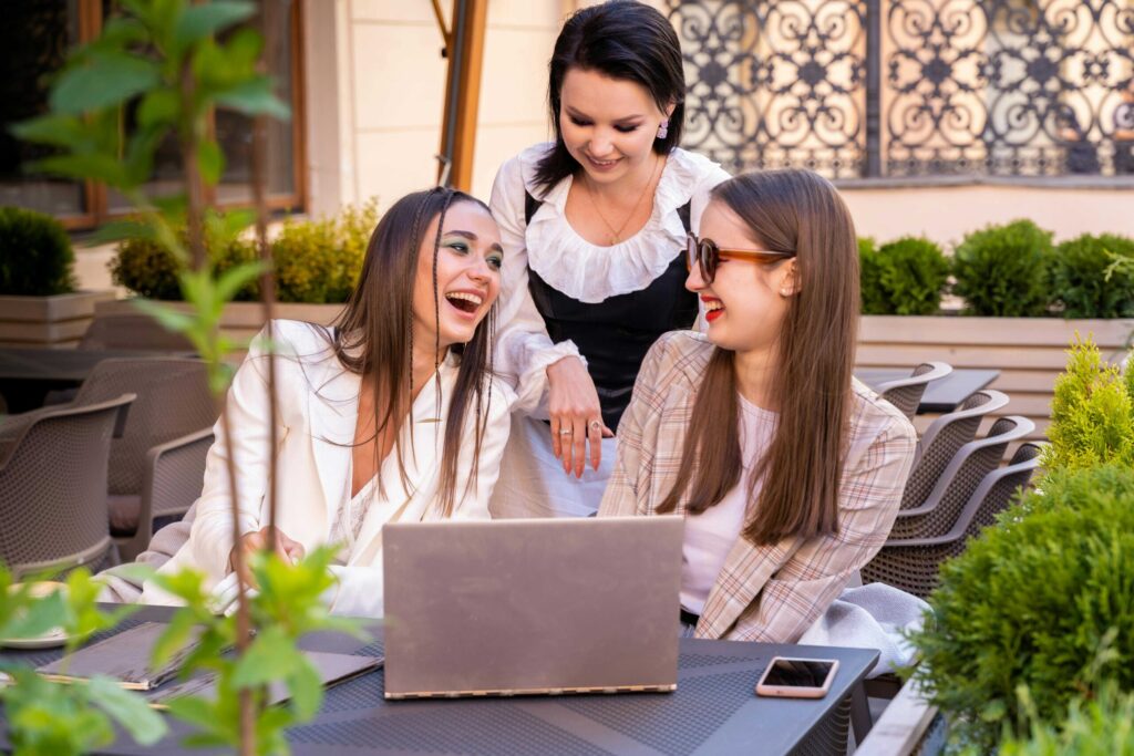 Three friends at an outdoor cafe smiling at a laptop screen, casually dressed and chatting.