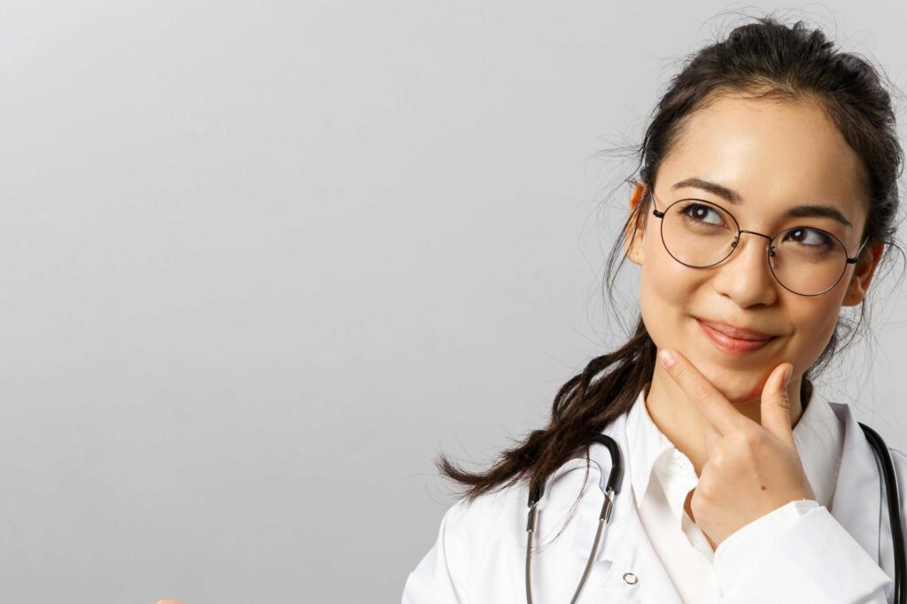Thoughtful female doctor wearing round glasses and a stethoscope, looking to the right with hand near chin in a white coat.
