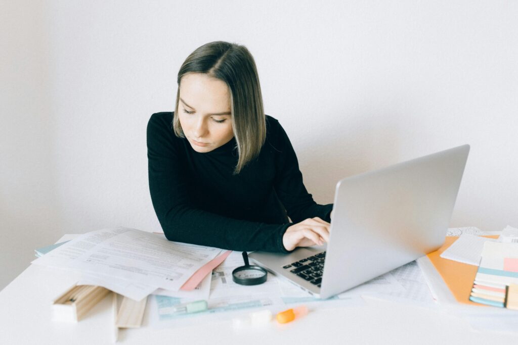 Woman in a black sweater working at a cluttered desk with papers, a laptop, and office supplies.