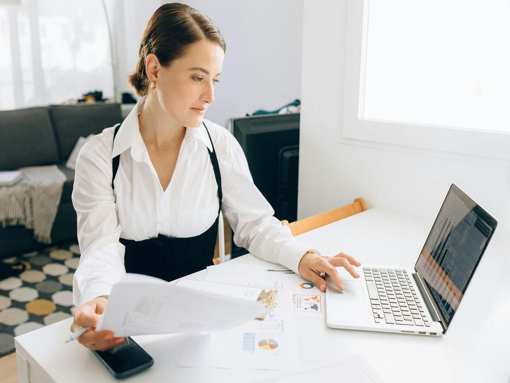 Professional woman at a white desk reviewing documents while using a laptop and phone nearby